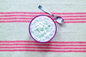 A bowl of greek yogurt dip, or tzatziki, with cucumber, dill and garlic. Shot from above on patterned linen with silver spoon.