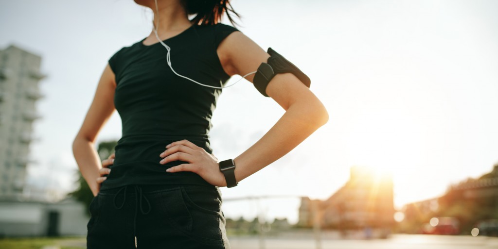 Cropped shot of urban runner outdoors in sunny morning. Fit young woman with smartwatch and mobile phone armband standing with her hands on hips.